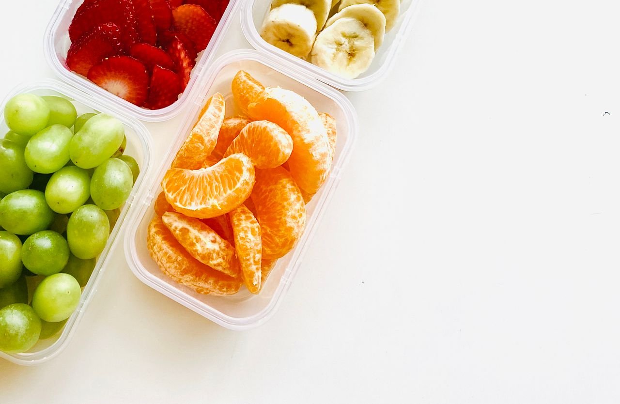 Top view of vibrant green grapes, orange slices, strawberries, and bananas in containers.