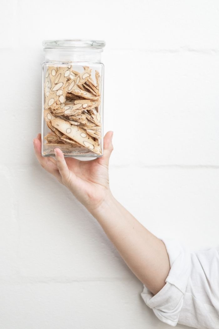 A hand holding a glass cookie jar filled with almond crisps against a white wall background.