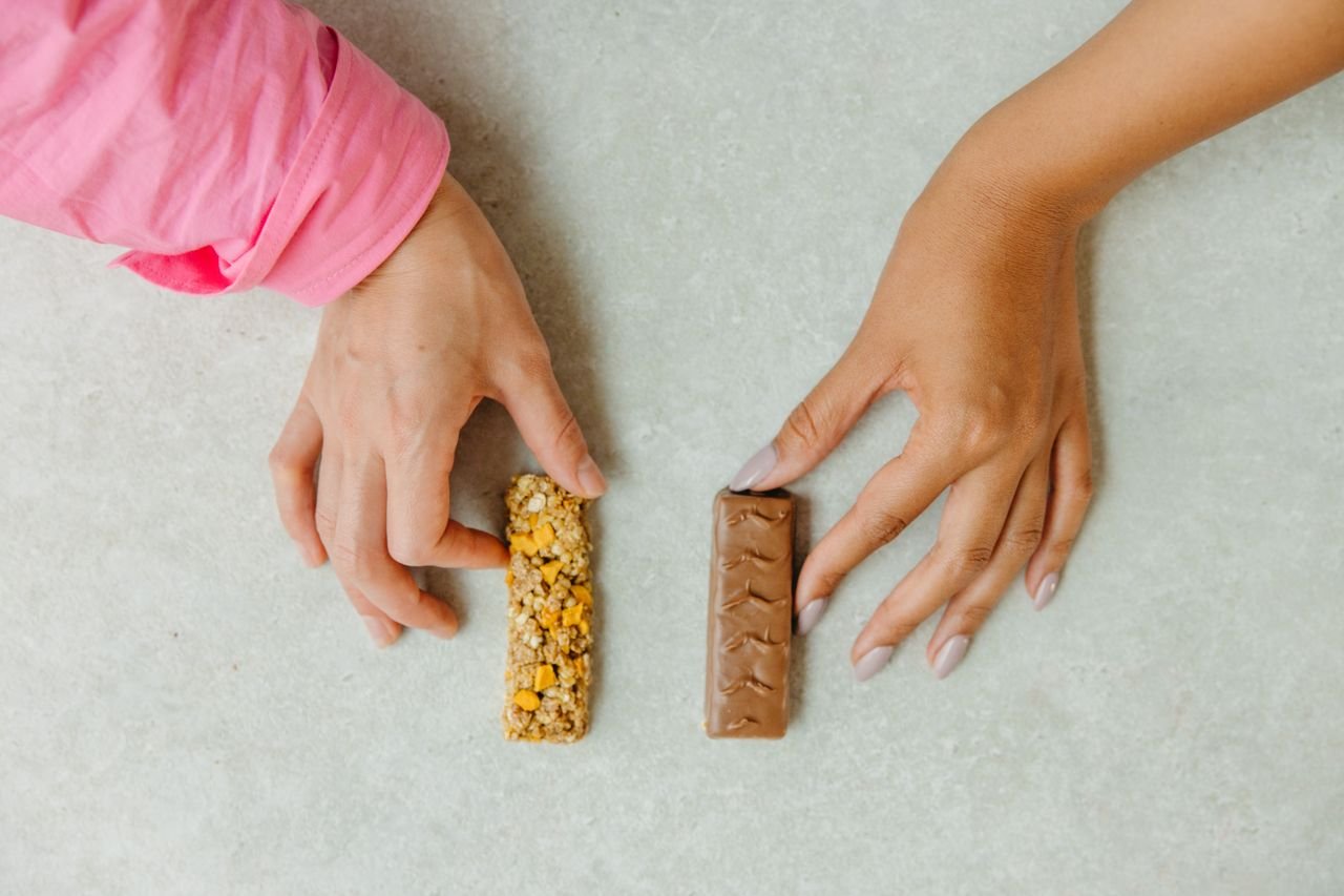 contact-img Two hands holding a chocolate bar and a granola bar, showcasing a snack choice concept.